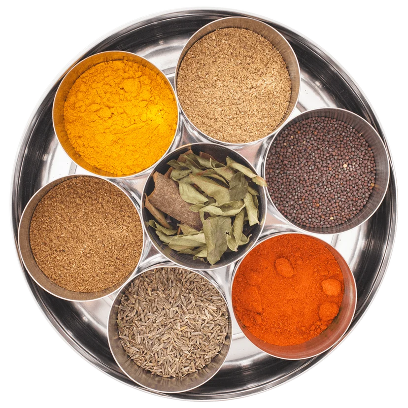 A stainless steel spice tray with six small metal bowls containing assorted whole and ground spices, including turmeric, cumin, mustard seeds, and coriander, arranged on a white background.