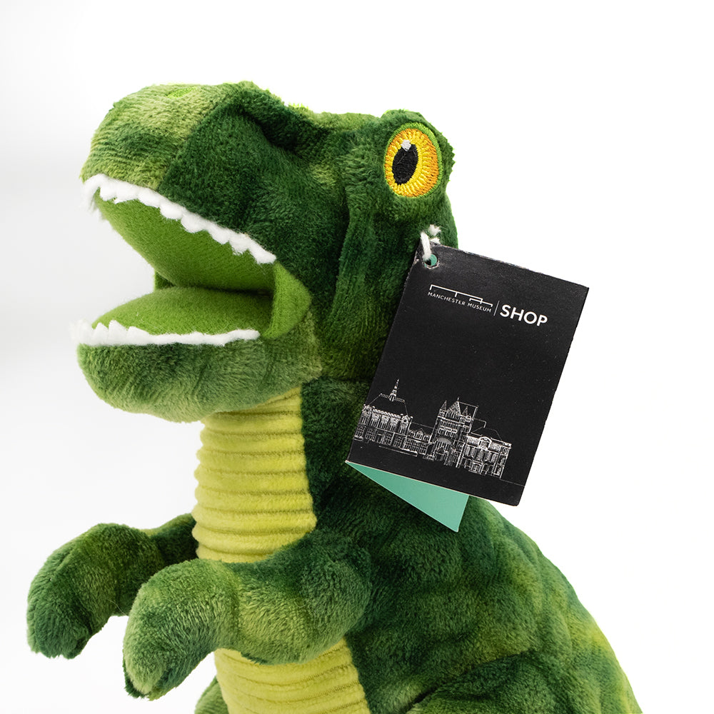 Close up image of a green fabric toy in the shape of a T-Rex pin front of a white background. Manchester Museum Shop label is attached.