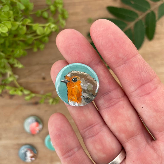 A person's hand holding a small, circular magnet featuring a hand-painted image of a robin. The robin is depicted in vibrant orange, brown, and grey colours, with a teal background. There are other similar magnets visible in the background.