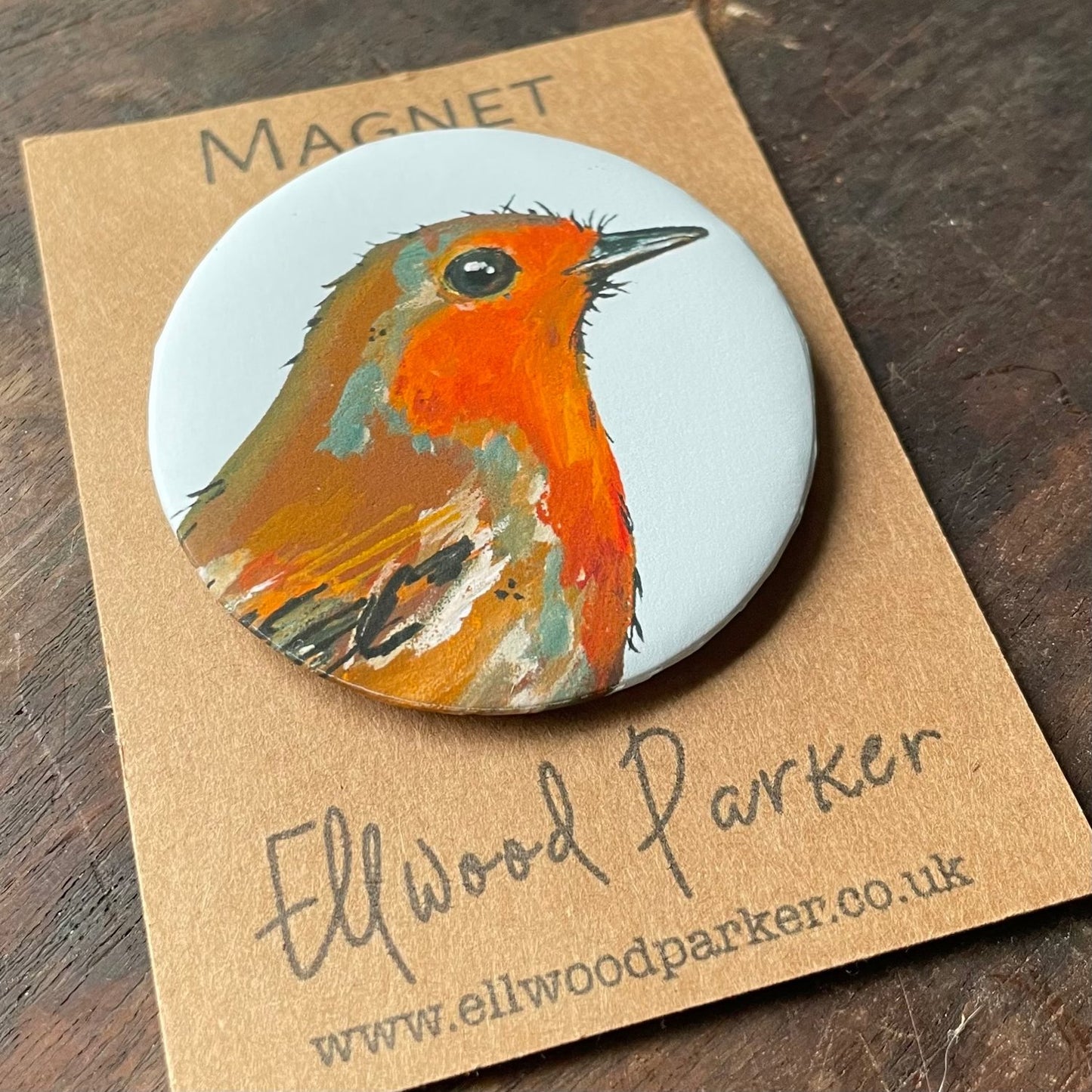 A circular magnet featuring a hand-painted image of a robin. The robin is depicted in vibrant orange, brown, and grey colours. The magnet is packaged in a brown cardboard sleeve with the artist's name.