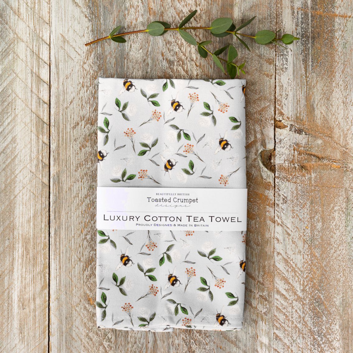 Tea towel with bee and floral pattern folded into a rectangular shape with a white belly band with Toasted Crumpet branding. Photographed on a wooden surface.