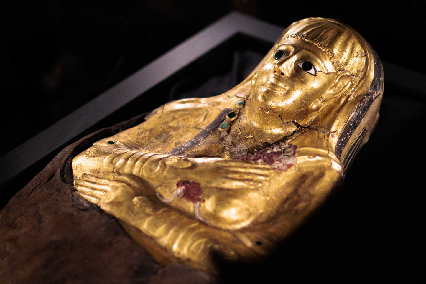 Golden mummy mask cartonage of a woman with her arms over her chest, the left arm is higher up and the right arm reaches straight across her body. Snake shaped bracelets are on her arms.