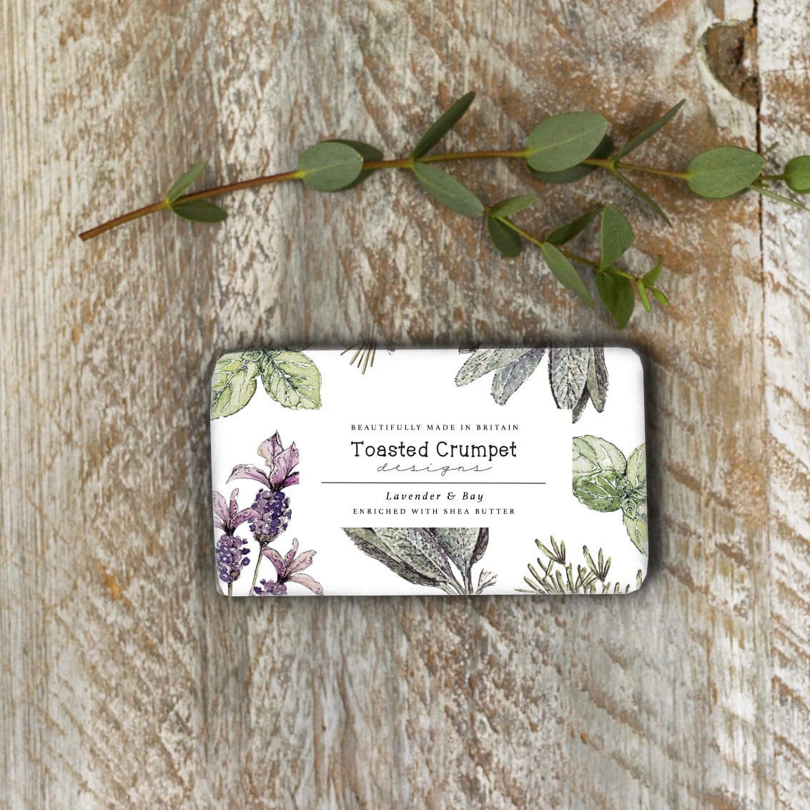 Lifestyle of rectangular soap with lavender flowers and herbs on the paper packaging. It's on a rustic white-washed surface and an eucalyptus twig is lying above the soap.