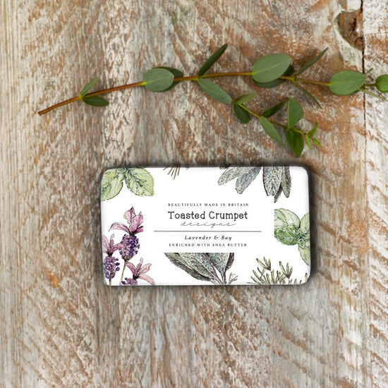 Lifestyle of rectangular soap with lavender flowers and herbs on the paper packaging. It's on a rustic white-washed surface and an eucalyptus twig is lying above the soap.