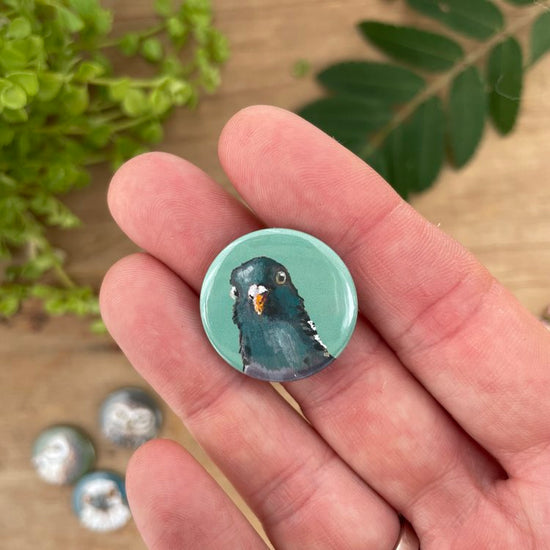 A person holding a circular pin badge with an illustration of a pigeon