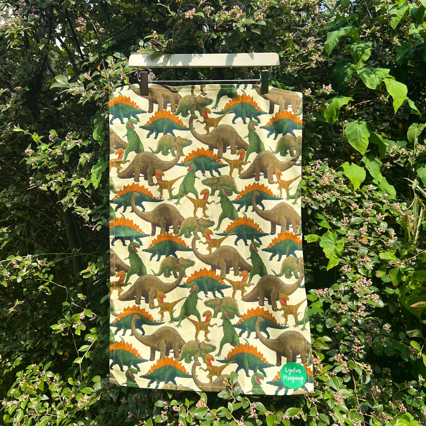 Tea towel with dinosaur repeat pattern hung up against a garden backdrop