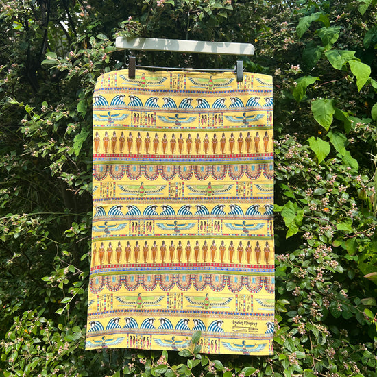 Tea towel with egyptian-inspired design hung on a hanger and held up against a garden backdrop.