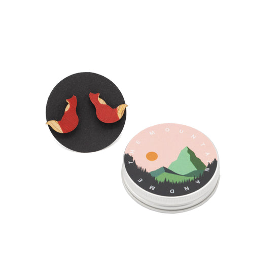 Earrings in the shape of a red fox against black backing card. The lid of a keepsake tin is to the right.