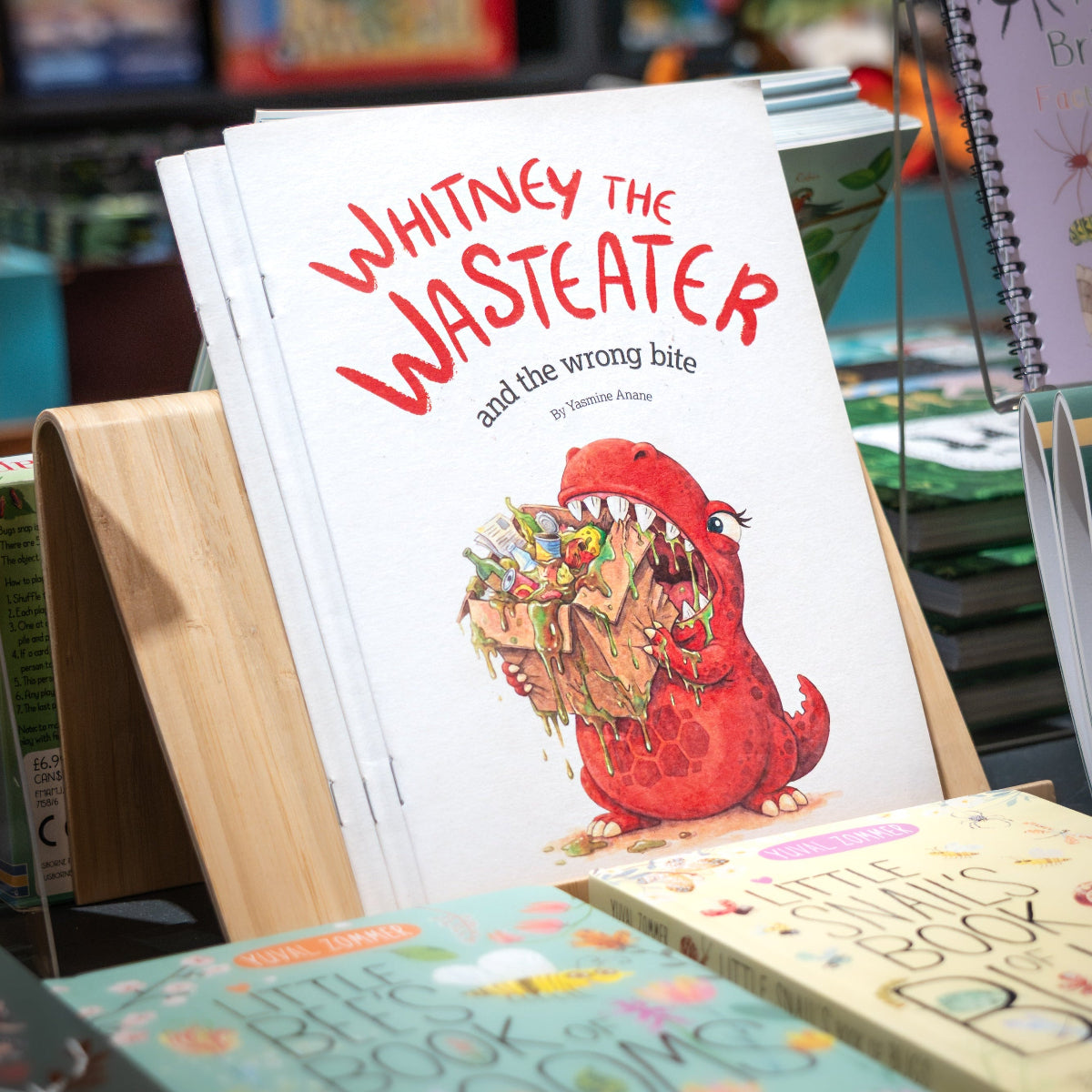 A children's book titled "Whitney the Wastemaker and the wrong bite" displayed on a wooden stand in a bookstore, with other books blurred in the background.