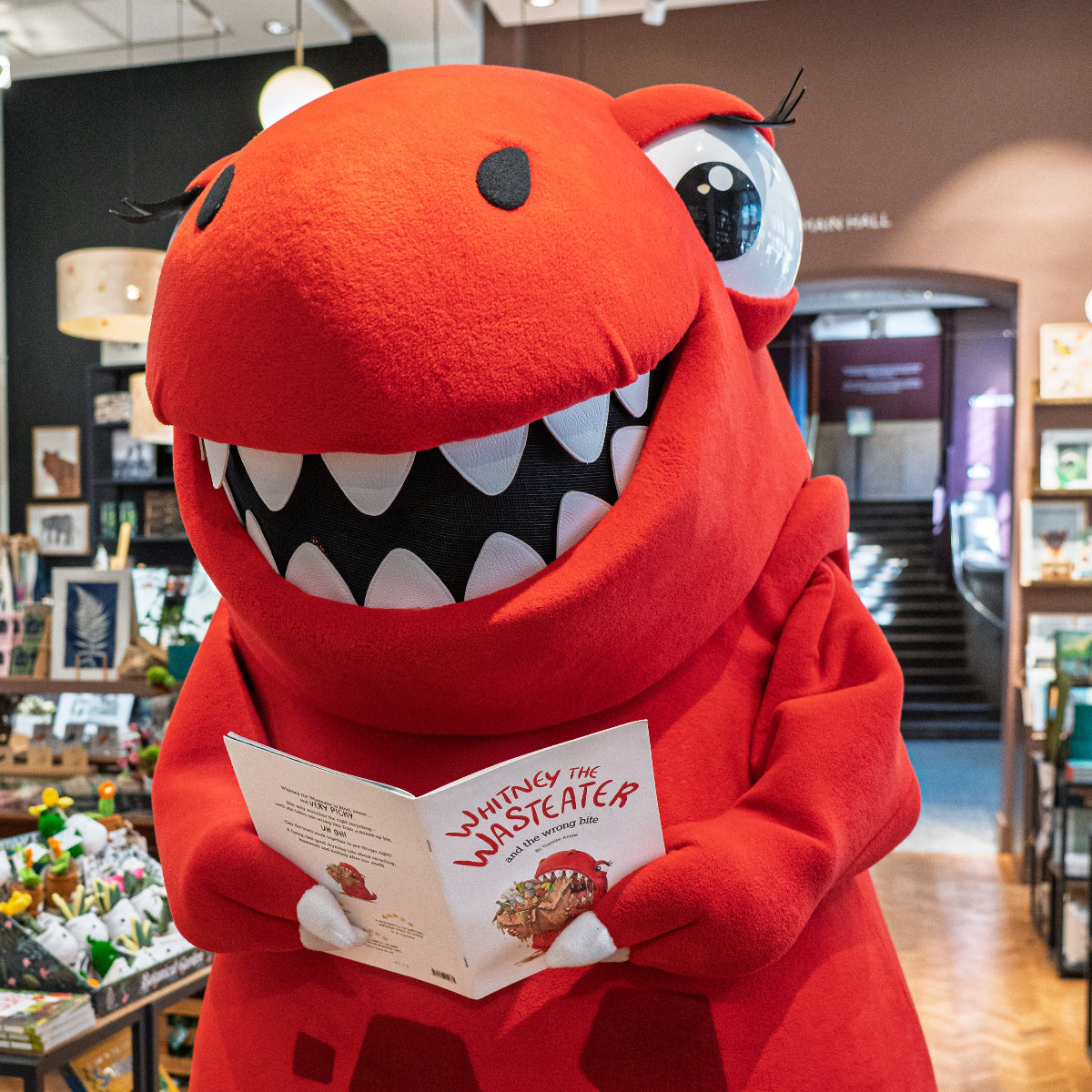 A red dinosaur mascot costume standing in a retail store, holding a children's book titled 'Whitney the Wasteater'. The plush costume features a wide grin with white teeth, large cartoon eyes, and a dark red hexagonal pattern on the belly.