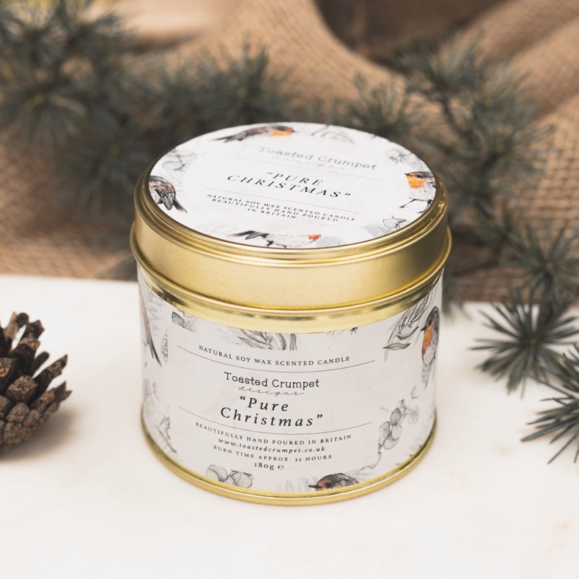 A tin candle with a gold lid and label that reads 'Pure Christmas', surrounded by decorative holiday elements.
