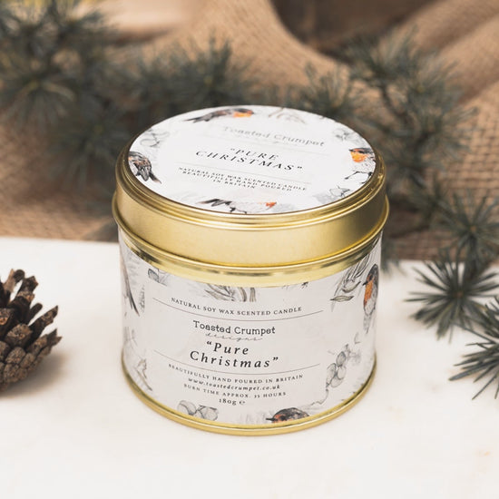 A tin candle with a gold lid and label that reads 'Pure Christmas', surrounded by decorative holiday elements.
