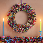 A multi-coloured wreath made out of recycled saris presented above a mantlepiece with two candles to the side.