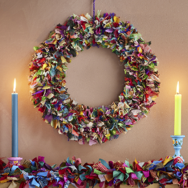 A multi-coloured wreath made out of recycled saris presented above a mantlepiece with two candles to the side.