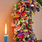 A close up of a multi-coloured wreath made from recycled saris with a lit candle to the left.