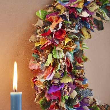 A close up of a multi-coloured wreath made from recycled saris with a lit candle to the left.