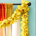 A yellow fabric garland hanging from a curtain rod. The garland is made of strips of recycled sari fabric in various patterns.