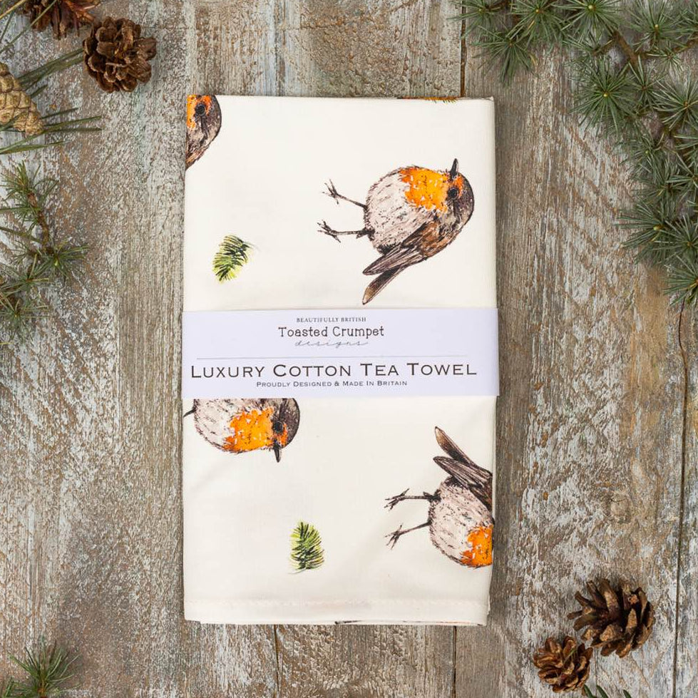A folded beige cotton tea towel with a printed robin design, presented on a wooden surface with pine cones and decorative tags.
