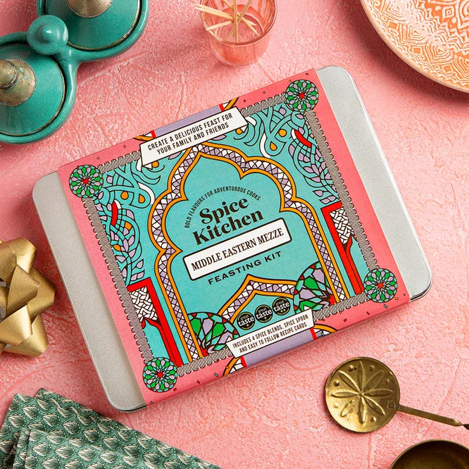 Middle Eastern Mezze Feasting Kit by Spice Kitchen in a decorative tin, photographed on a pink textured surface with colourful tableware and serving utensils.