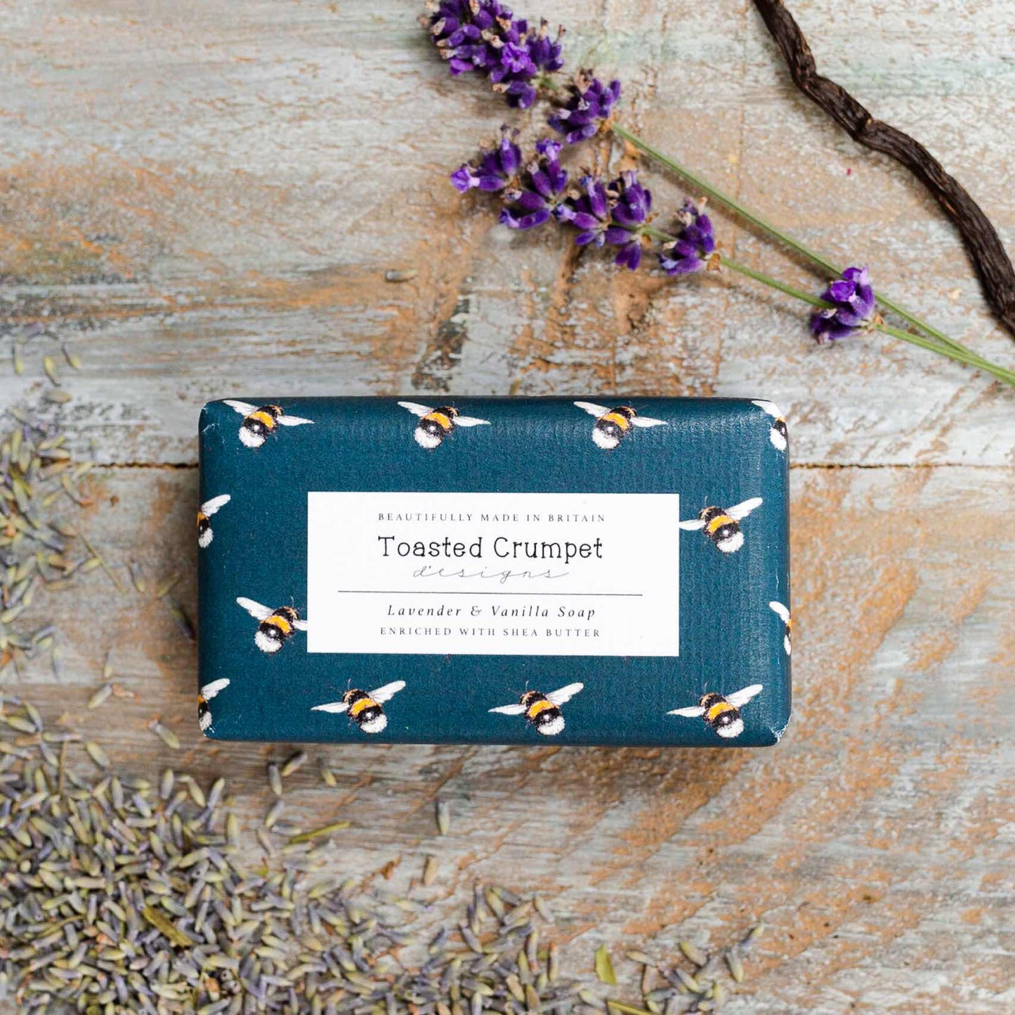 Rectangular soap wrapped in bee-patterned packaging with a label by Toasted Crumpet.  Photographed against a wooden surface with some lavender in the corner.