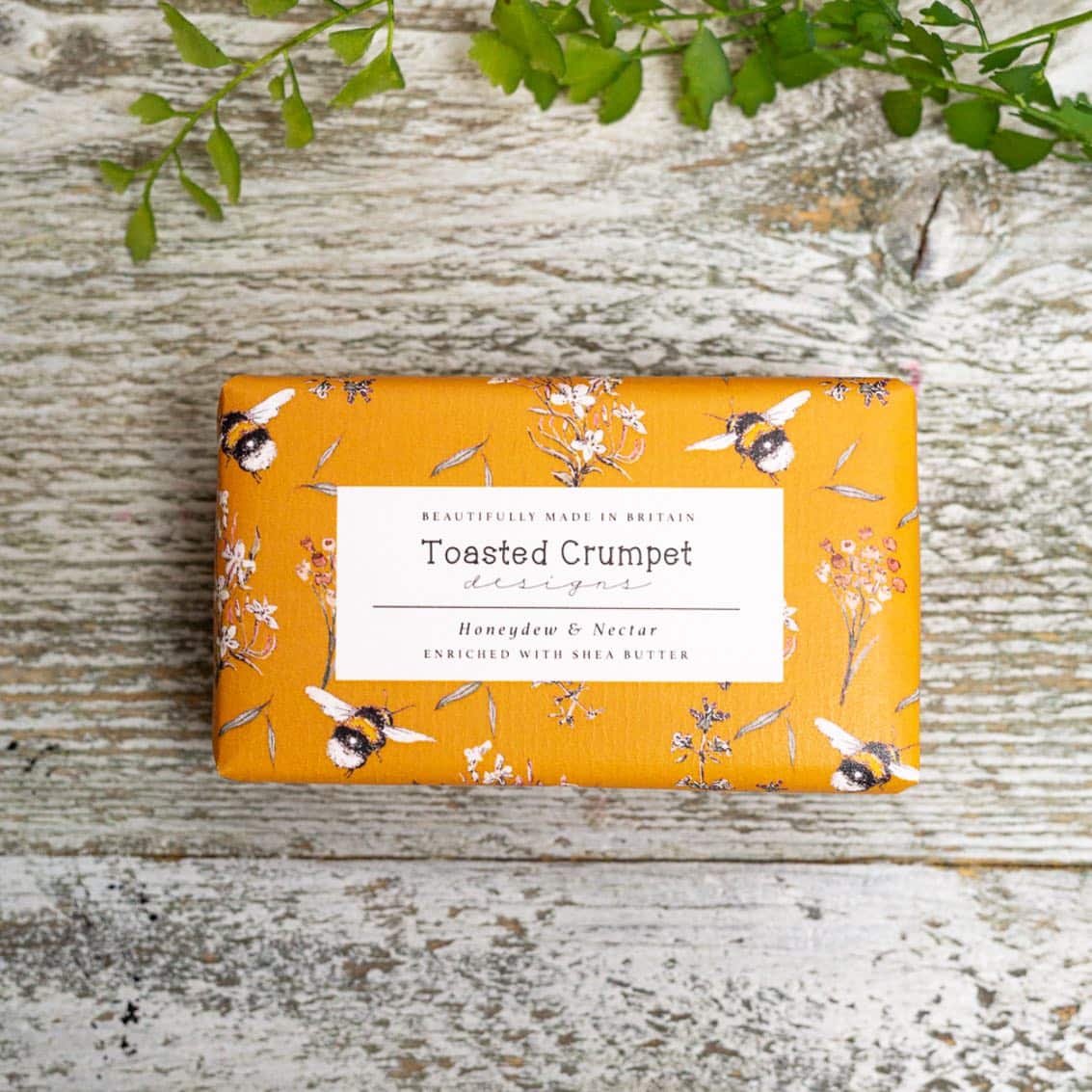 Rectangular soap wrapped in orange packaging with pictures of bees. Photographed birds-eye view on a wooden surface.