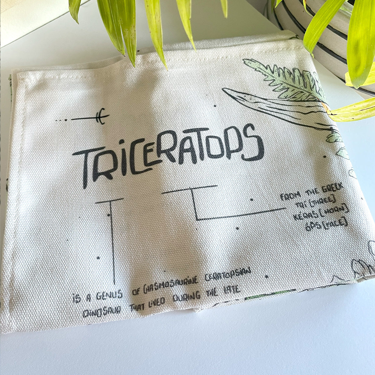 Close up of tea towel with 'Triceratops' design and text on a white surface with green leaves.