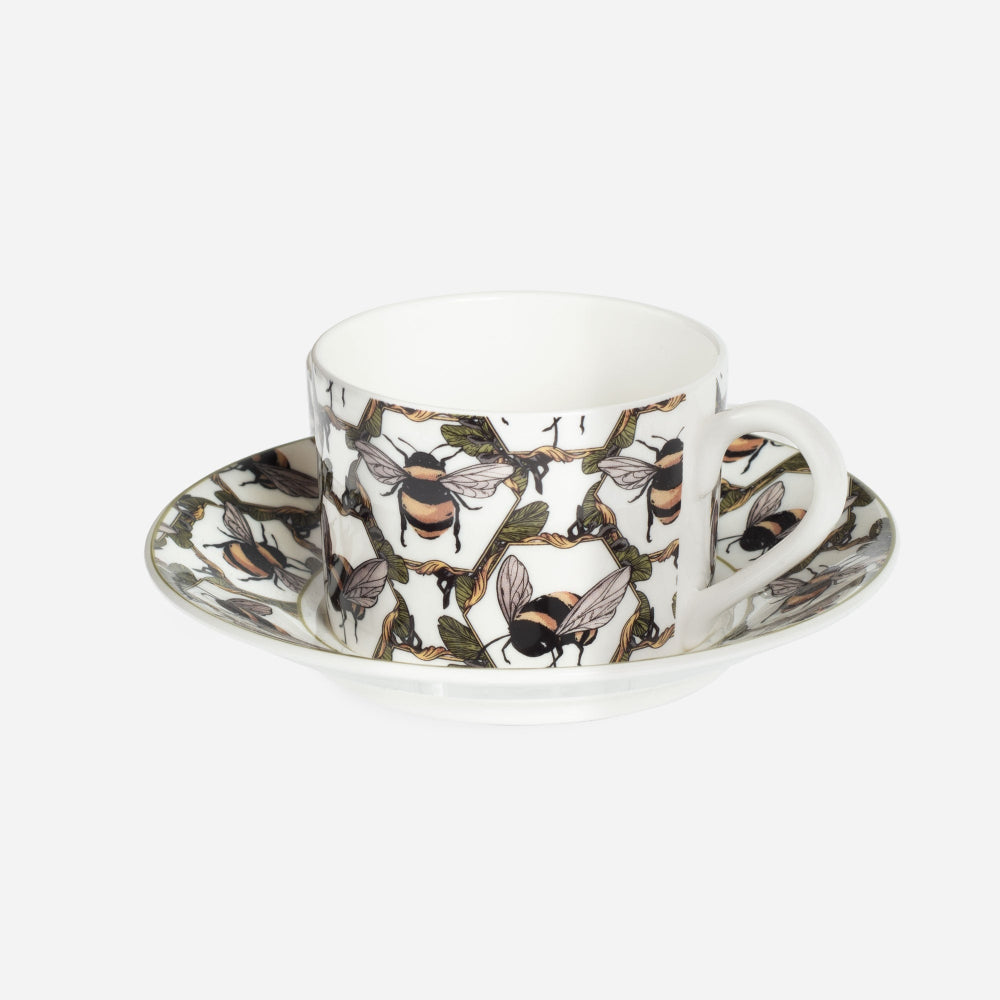 A white ceramic teacup and saucer set decorated with an all-over illustration of bees and a honeycomb pattern, shown on a light gray background.