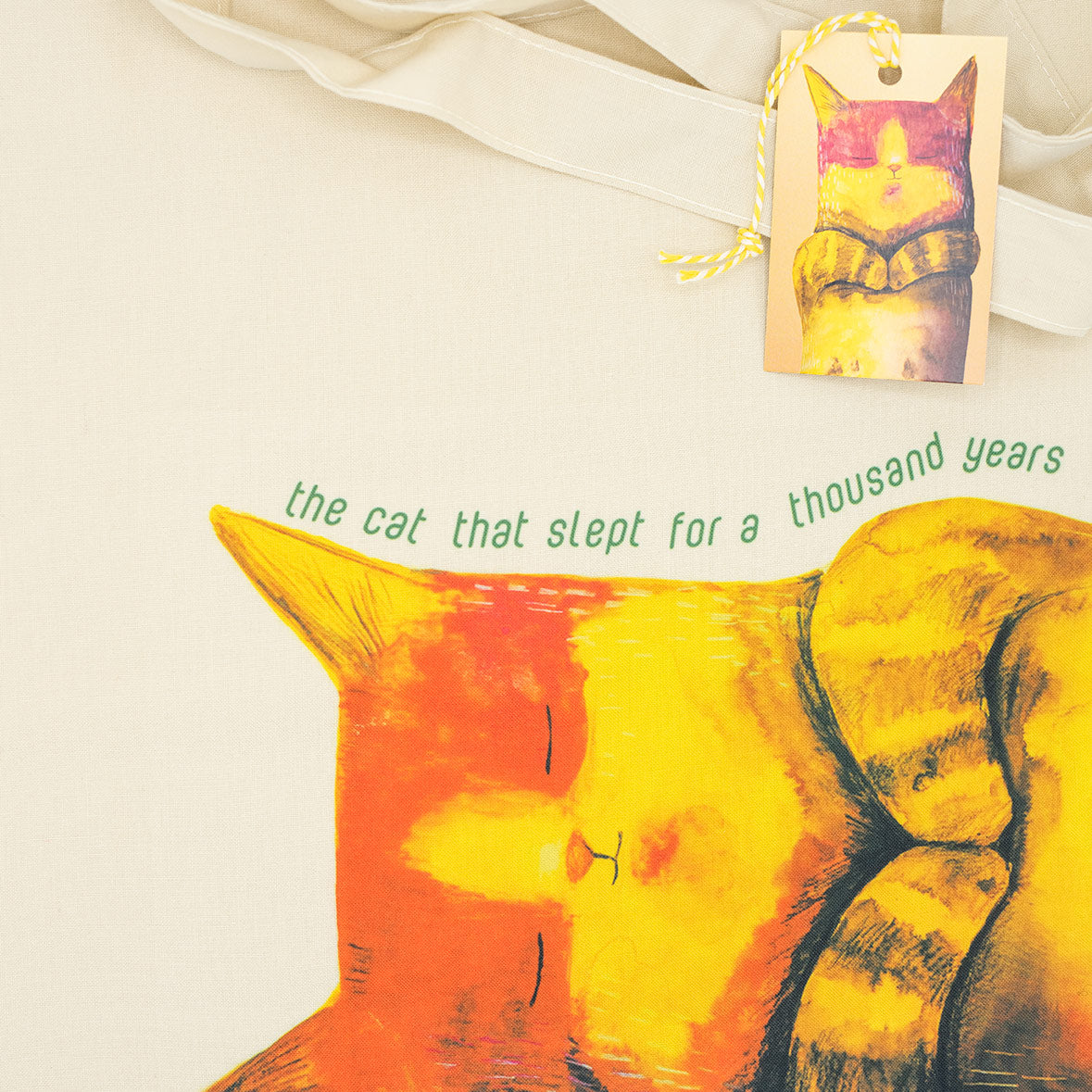 Close up of a tote bag with an illustration of a cat on it with text. The label with the same illustration is also placed.