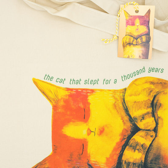 Close up of a tote bag with an illustration of a cat on it with text. The label with the same illustration is also placed.