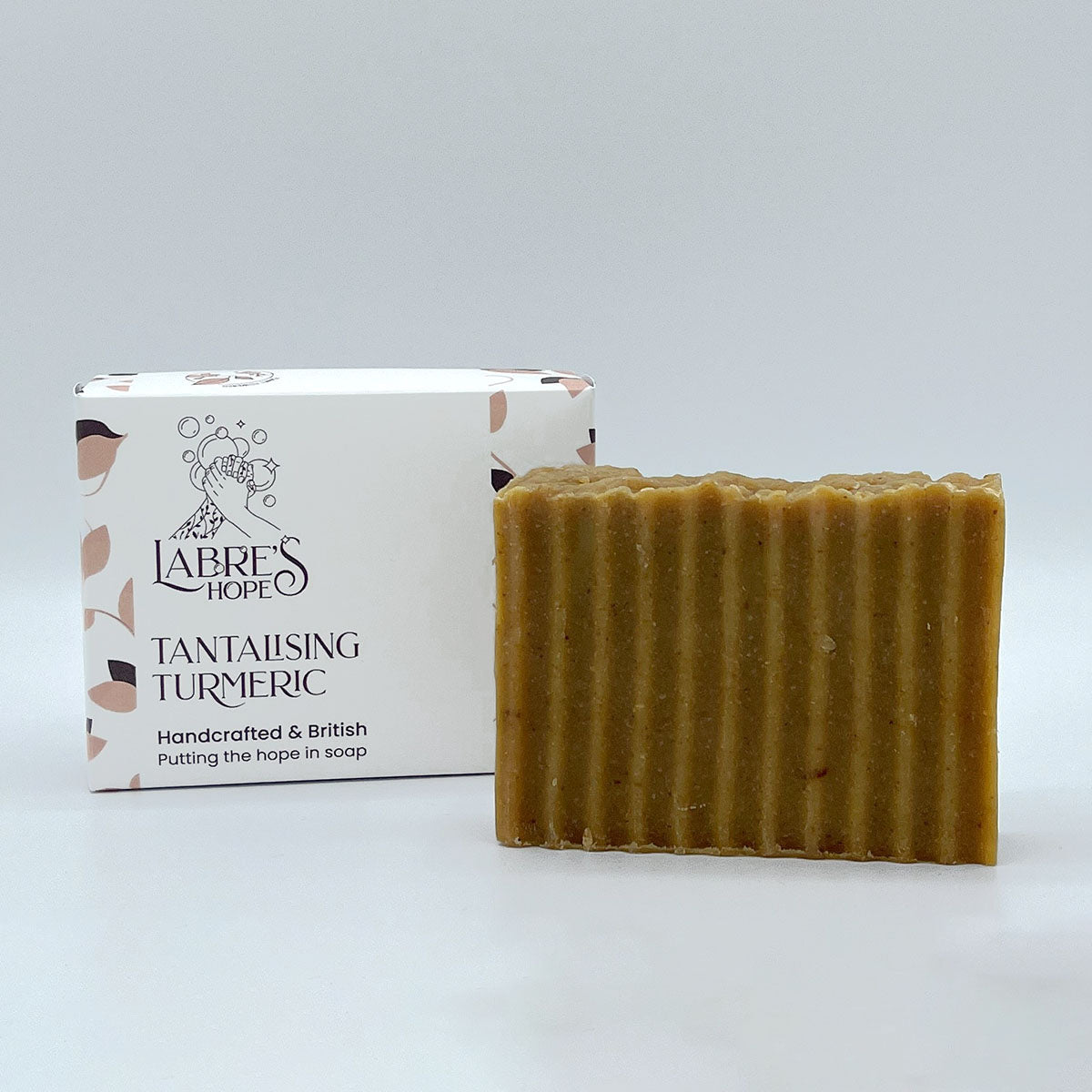 A rectangular bar of soap with a golden brown colour. The soap is resting inside a white cardboard box with a black and white label. 