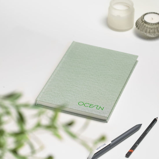 Lifestyle of green notebook with the text: OCEAN on the front. A pen and a pencil are to the bottom right..