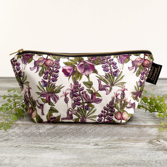 Rectangular cosmetics bag with white, purple and green floral pattern design, photographed upright on a grey wooden surface. Bag has 2 Toasted Crumpet branded. labels attatched.