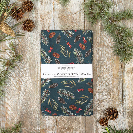 A navy-colored cotton tea towel with a Christmas-themed pattern of pine cones and red berries, labeled 'Toasted Crumpet Luxury Cotton Tea Towel', displayed on a wooden surface with pine branches around.