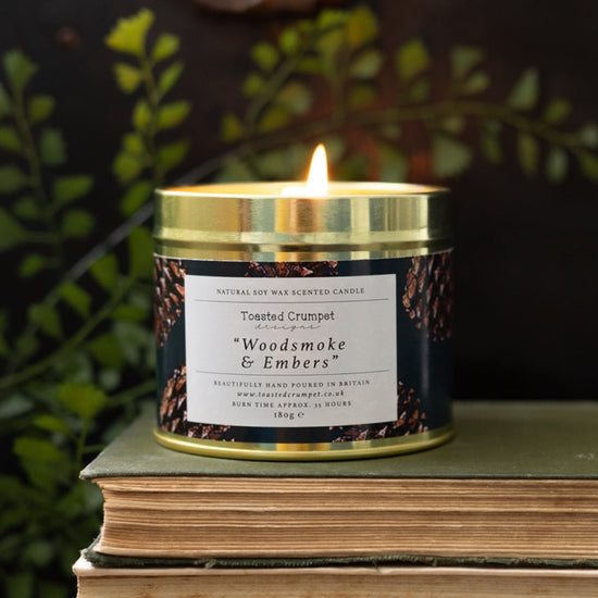 A lit candle in a tin container with a gold lid, placed on a book, with the label 'Woodsmoke & Embers'. The candle is surrounded by green foliage.