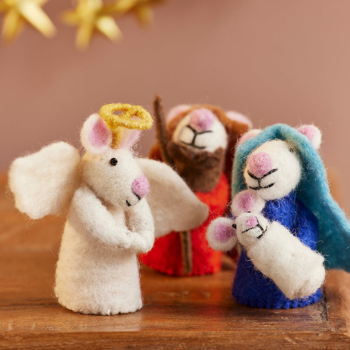 Handmade felt wool Christmas mice nativity figurines on a wooden table, featuring an angel in white with a gold halo, a robed figure in red, and Mary in blue holding baby Jesus.