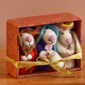 A small red gift box with a gold ribbon bow, containing four soft felted wool mice figures in different coloured outfits, displayed on a wooden surface against a warm beige background.