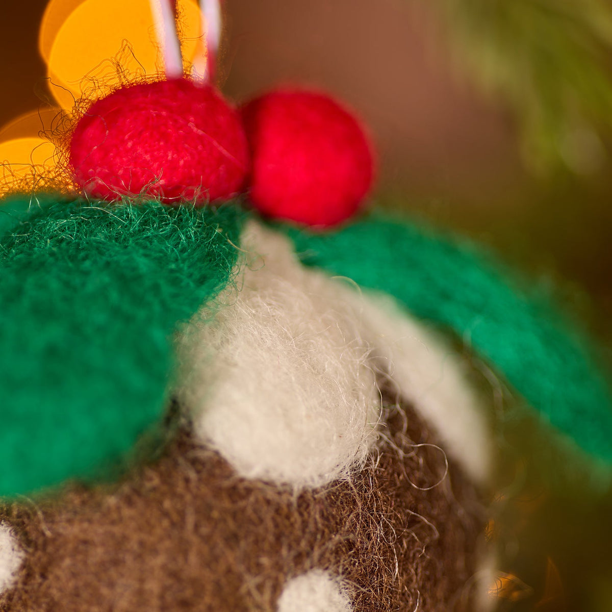 Close-up of a felt Christmas ornament shaped like a pudding, with a brown body, white polka dots, green leaves, and red berries, hanging against a blurred festive background with warm lights.