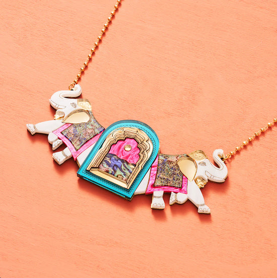 An archway pendant with a white elephant walking out from behind either side of it. The elephants have raised trunks and wear decorative cloth on their backs. the archway itself is turquoise around the edges with a wooden trim and pink and colourful centre. The gold chain is fastened to the elephants' raised trunks. Peach background.