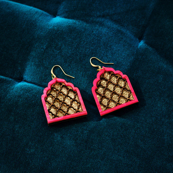 A pair of pink edged Jali-inspired earrings shaped like an arch with several curves. An internal lattice structure is created with wood on a gold background. Dark teal fabric backdrop.