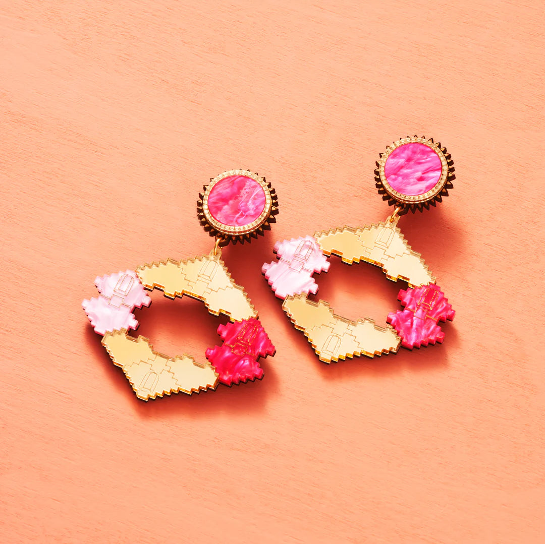 A pair of jagged edged diamond shaped earrings in gold, hot pink and pale pink. The stud part of the earringshas a round pink disc to hide the stud. Pale peachy backdrop.