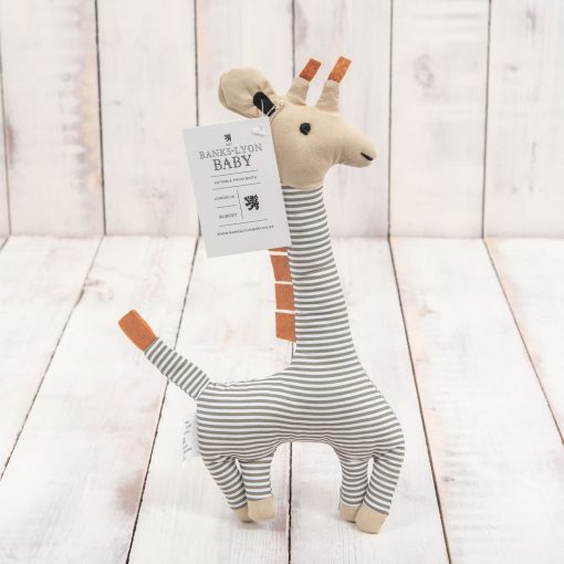 The giraffe soft toy standing on white stained wooden boards. The body is white and faded blue striped while the head is beige. The swing tag hangs from the ears.