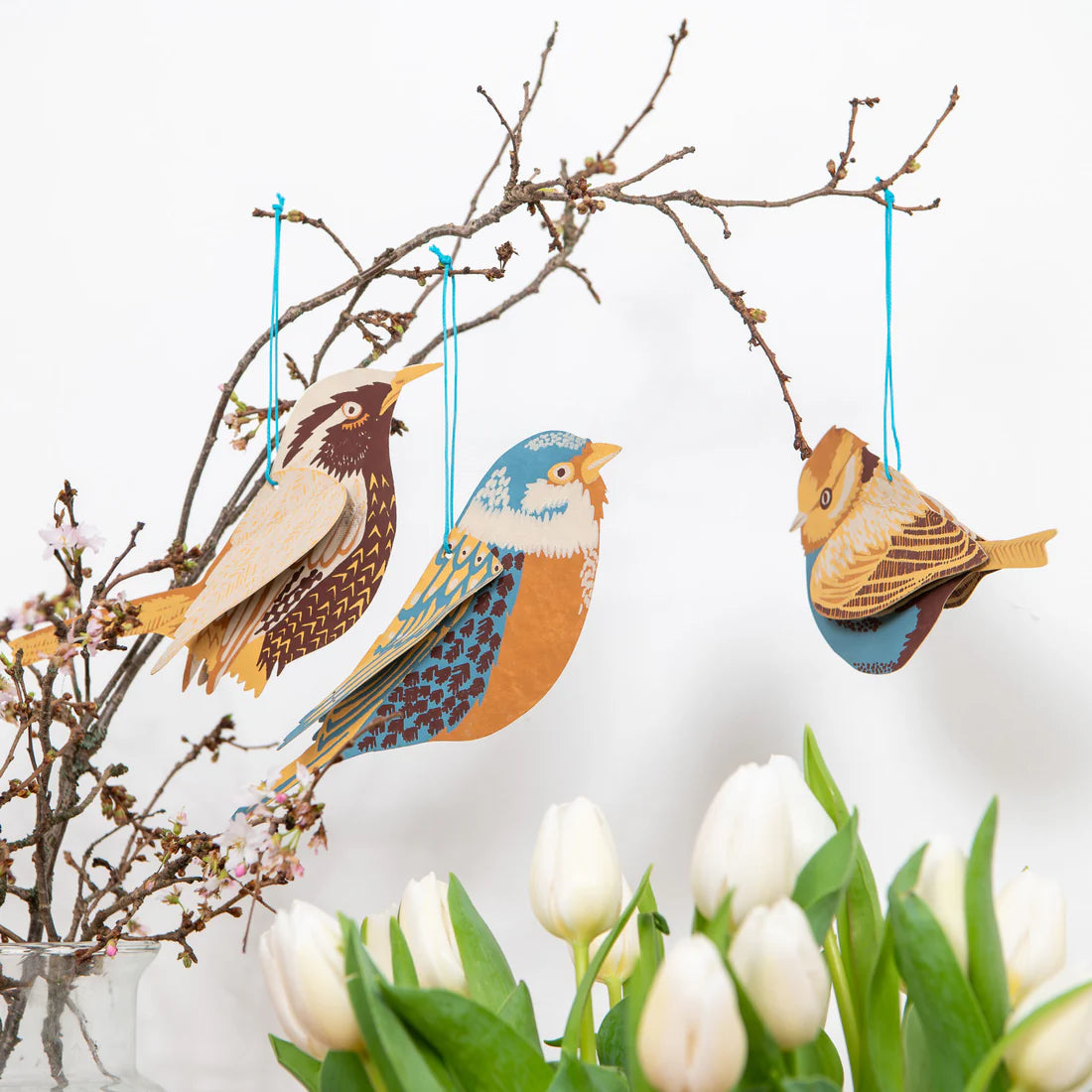 Three paper bird decorations on a branch hanging above white tulips.