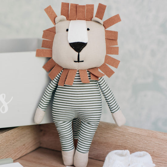 The lion soft toy standing up against a wooden and grey backdrop. The body is striped white and faded blue while the head and paws are beige. A brown felt mane circles the face.