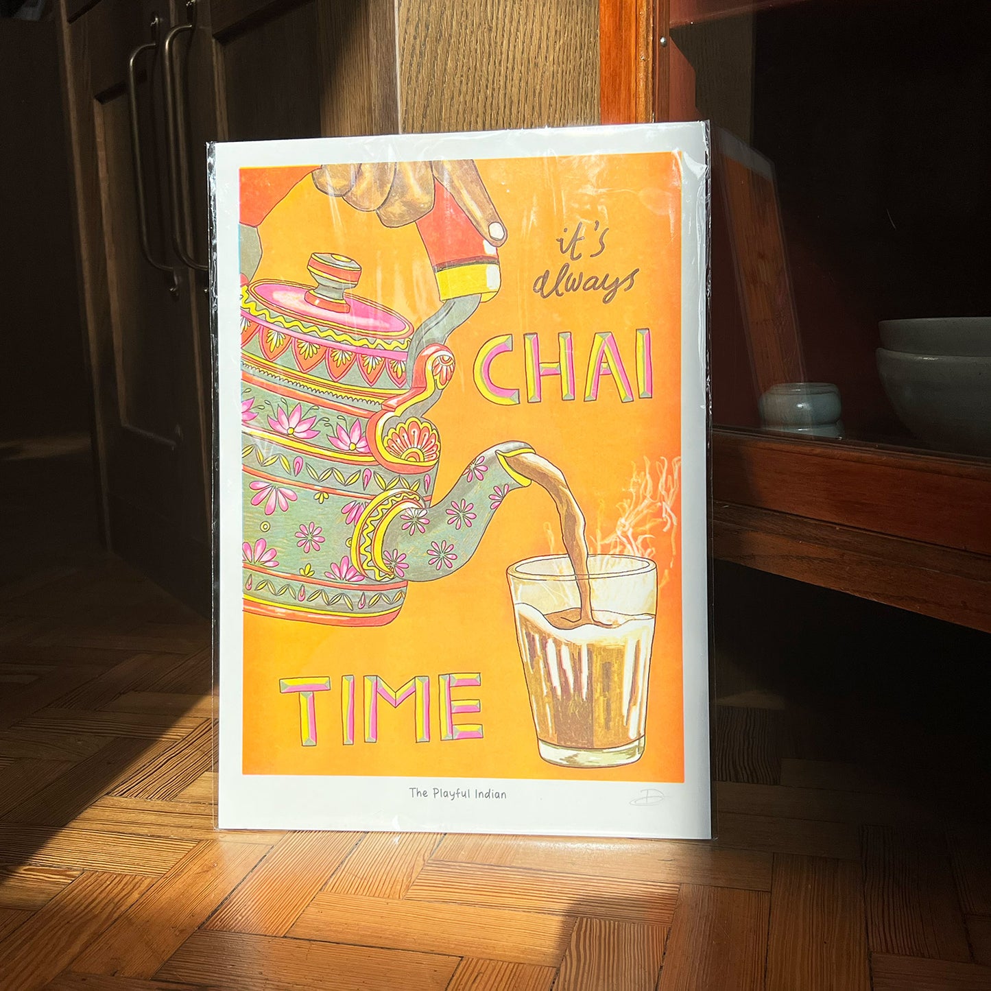 This bright and colourful print shows someone pouring a hot drink, called chai, from a pretty teapot into a glass, with the words "it's always CHAI TIME" written on it.