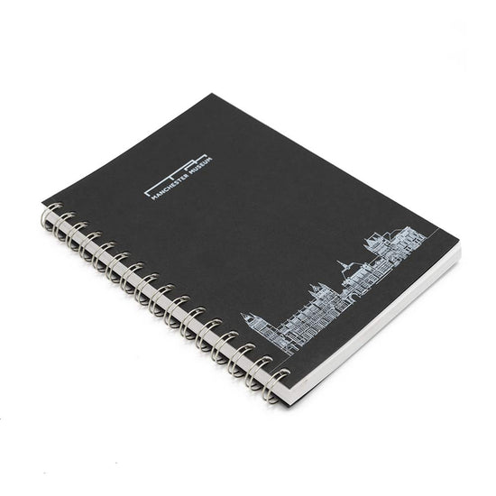 Wirobound black notebook photographed against a white background from a side angle view. The notebook has Manchester Museum's logo at the top centre in silver, and a silver line drawing of Manchester Museum at the bottom centre.