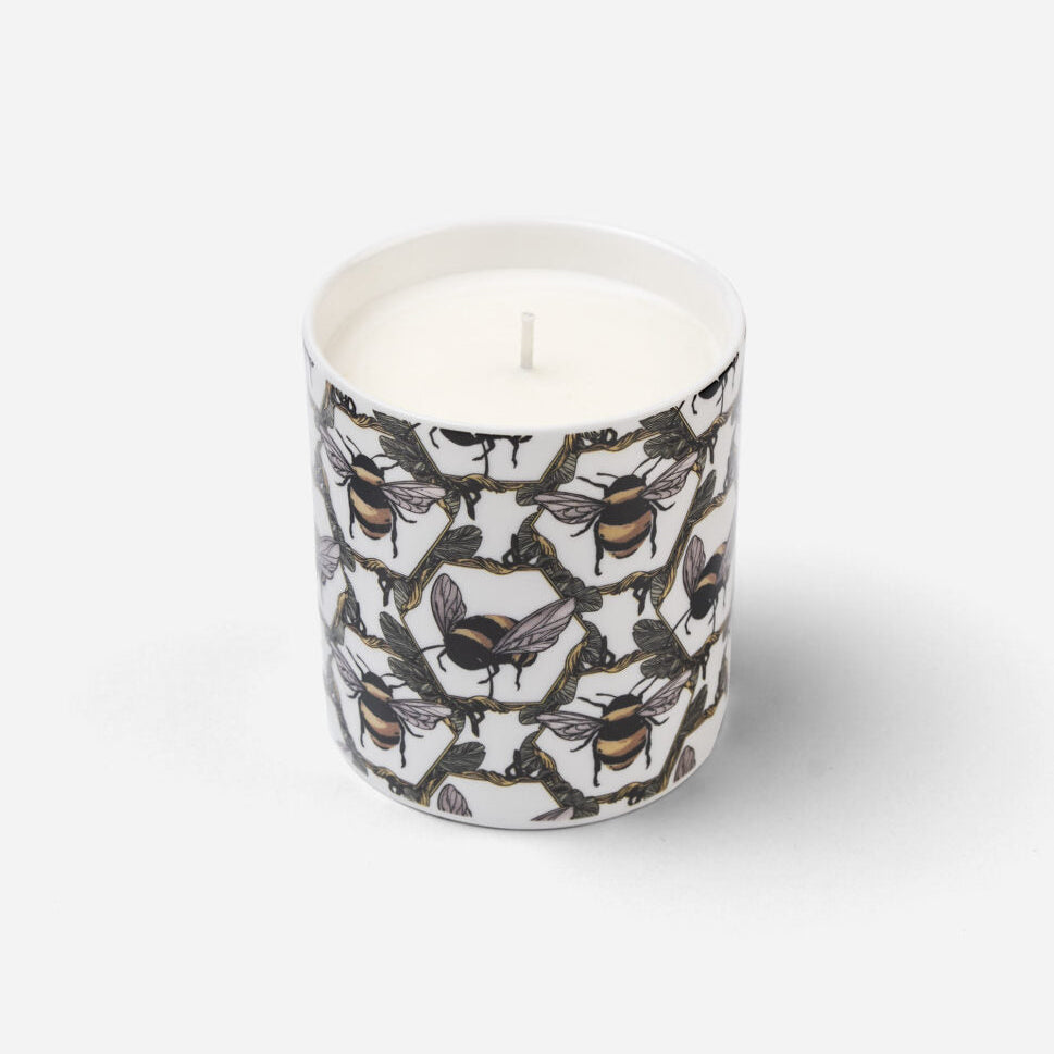 A white wax candle in a cylindrical container with an all-over pattern of bumblebees and honeycomb, set against a plain light gray background.