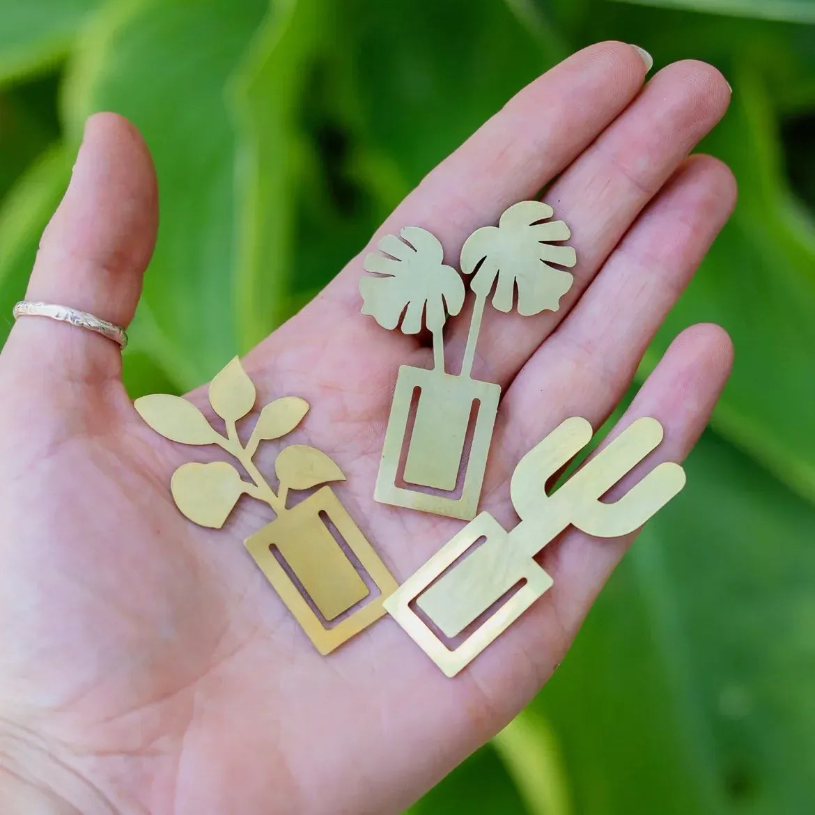 The image shows a hand holding three gold bookmarks shaped like different houseplants: a rubber plant, a monstera, and a cactus.