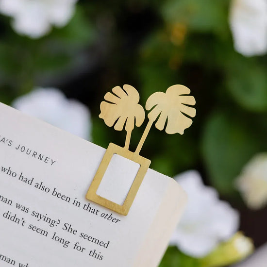 The image shows a gold bookmark shaped like a monstera plant, marking a page in a book.