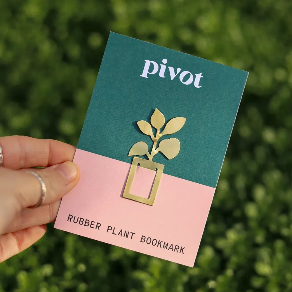 The image shows a hand holding a gold bookmark shaped like a rubber plant in a pot, with the word "PIVOT" printed on a card behind it.