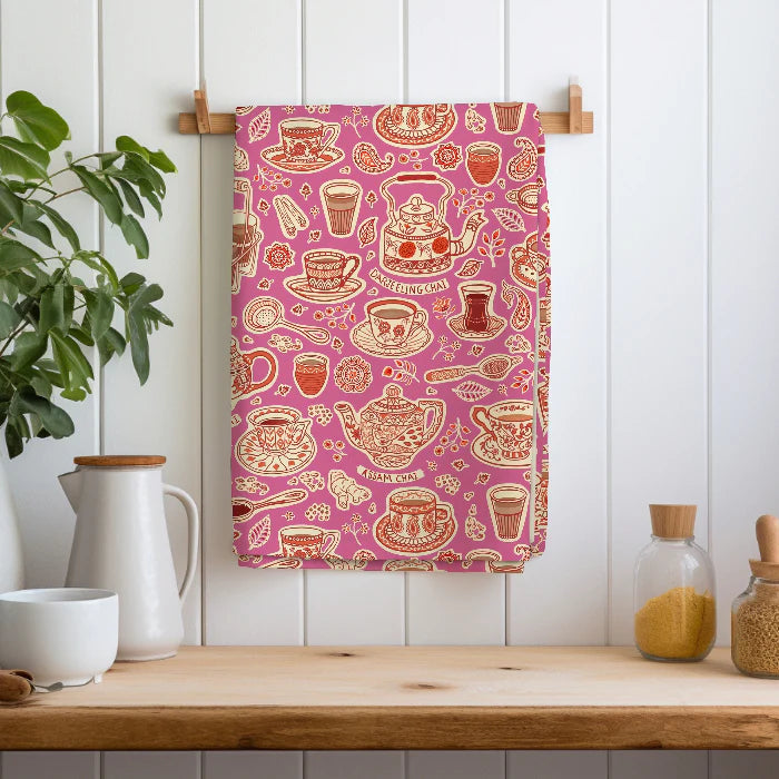 Tea-themed tea towel with teapots, cups, and spiceson a pink background hanging on a wooden rack.